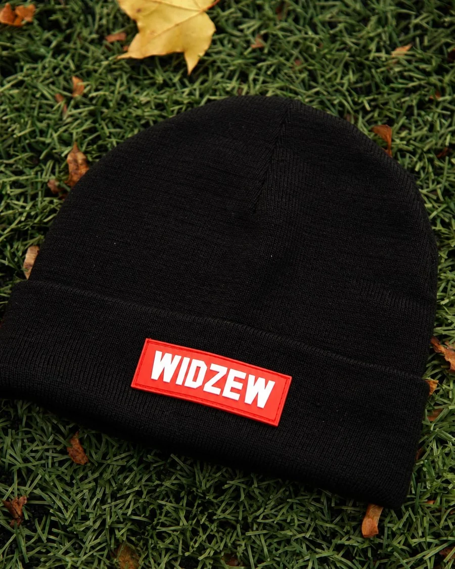 Czarna czapka beanie z wywiniętym mankietem i czerwoną naszywką z napisem Widzew, leżąca na trawie