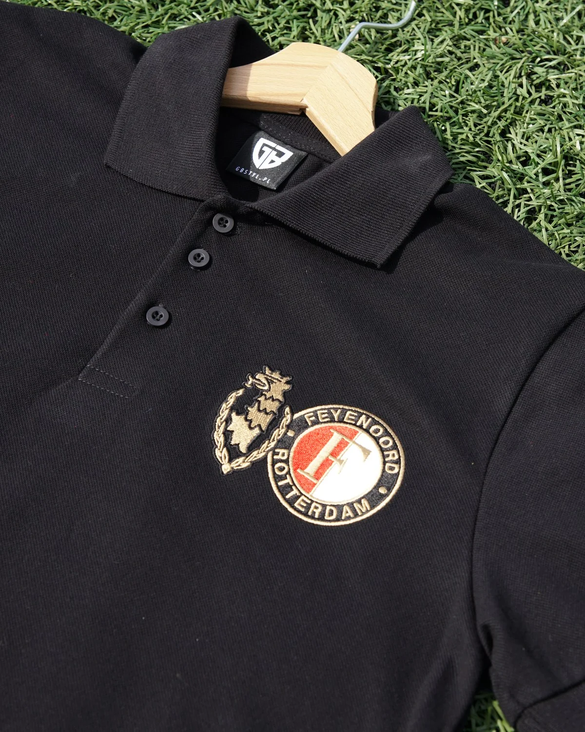 Zbliżenie czarnego polo na wieszaku, z herbem Feyenoordu Rotterdam i złotym znakiem na piersi