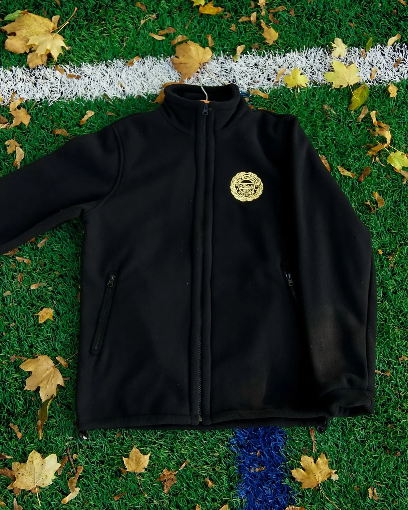 Black full-zip fleece with a stand collar and a gold embroidered crest on the chest, lying on grass
