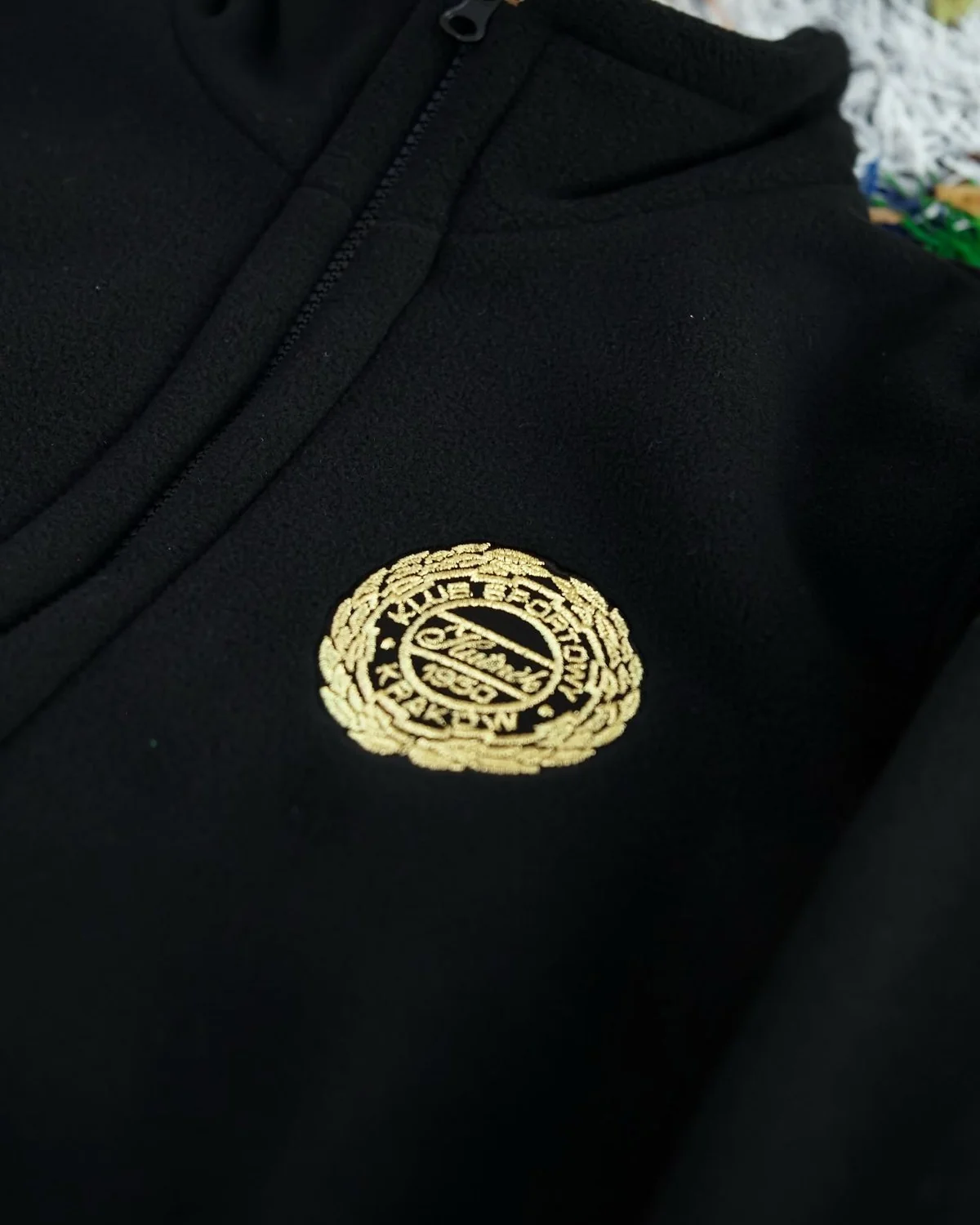 Close-up of the gold embroidered crest on a black fleece, next to the zip and stand collar