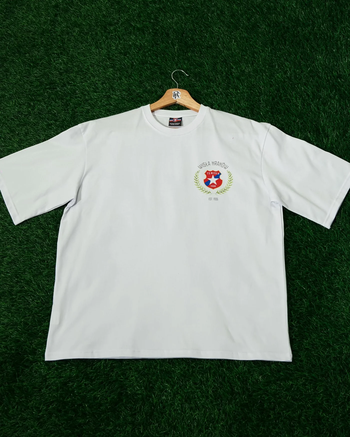 Biały t-shirt oversize na wieszaku, z niewielkim kolorowym herbem klubowym na piersi